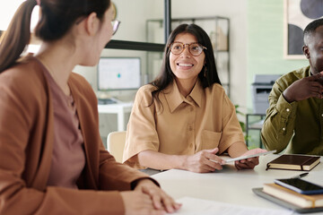 Smiling woman wearing glasses engaging colleagues at meeting table in modern office with large...