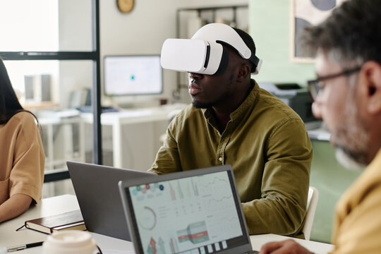Man immersed in virtual reality using VR headset while sitting in contemporary office space alongside two colleagues collaborating on digital devices