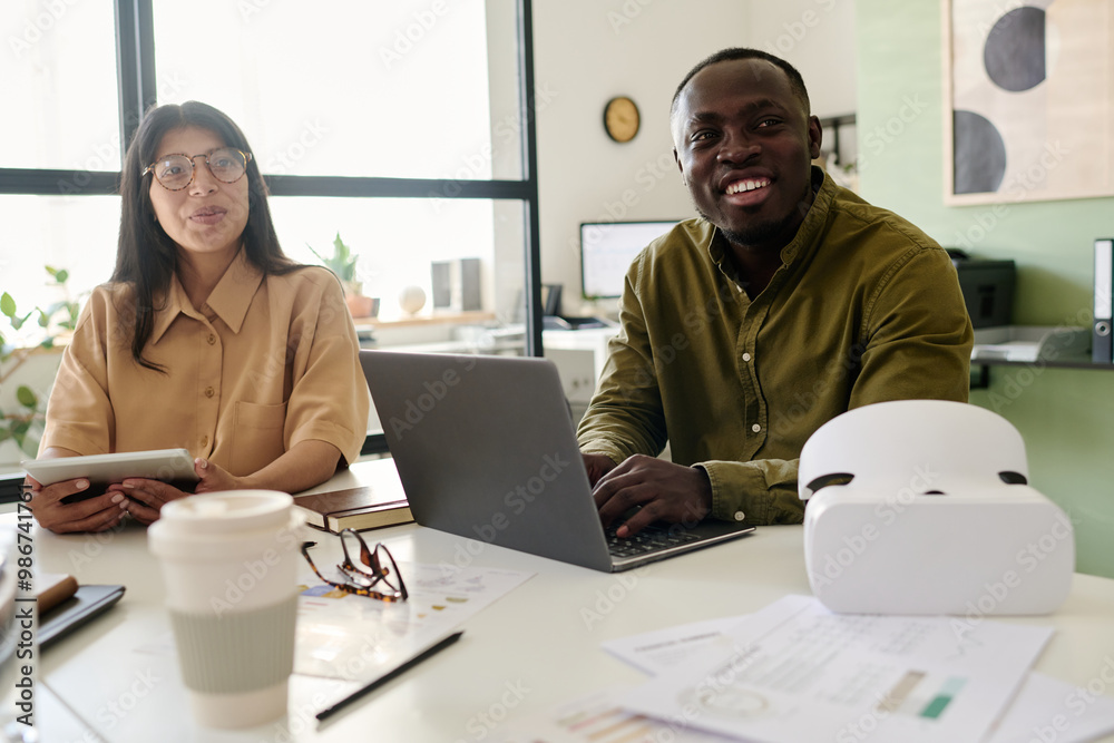 Poster Two colleagues, one Black person and one Caucasian, smiling and collaborating on project with laptop and documents on table, enjoying bright and open workspace
