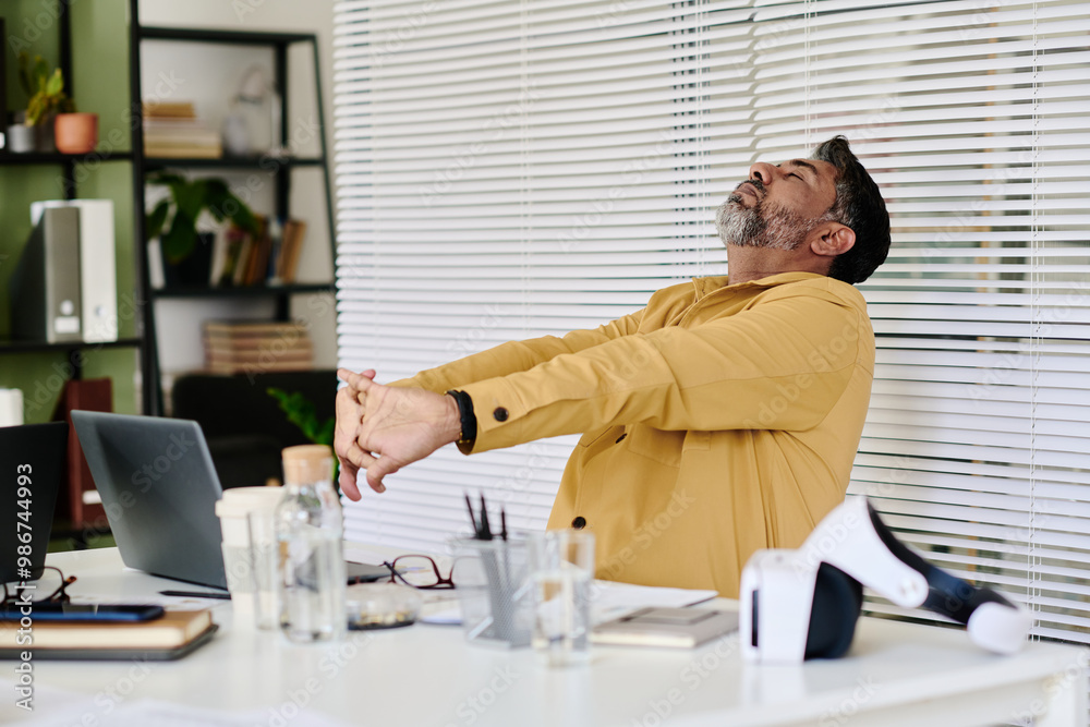 Poster middle-aged man stretching arms while sitting at desk in modern office setting with bookshelves and 