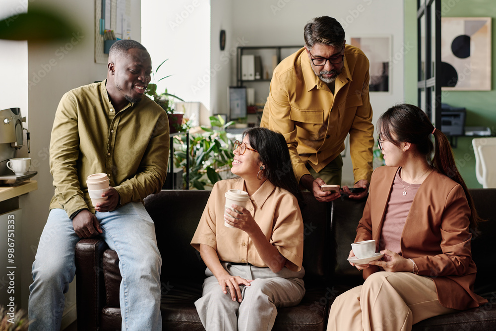 Aufkleber Group of diverse colleagues enjoying conversation while holding coffee cups in a modern office. Relaxed environment with comfortable seating and greenery around, fostering casual interaction