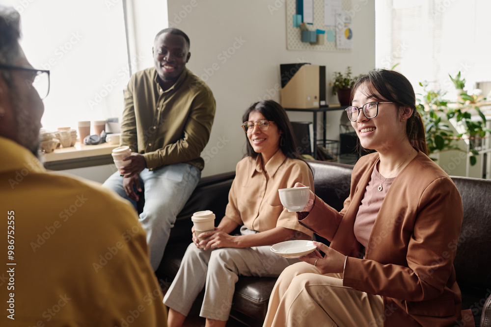 Poster group of diverse colleagues sitting on couch, smiling and holding coffee cups in bright office setti