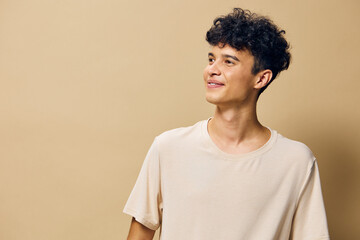 Young man with curly hair smiling against a warm beige background, wearing a casual light colored t...