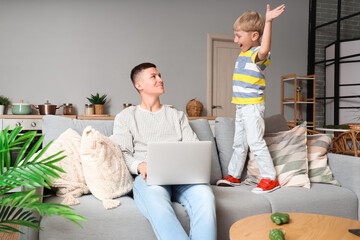 Working father with laptop and his naughty little son on sofa at home