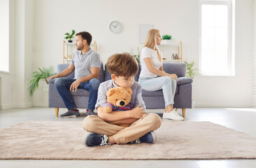 Frustrated upset boy son sitting on floor with toy bear while his mother and father sitting on...