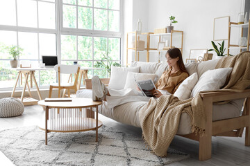 Pretty young woman reading magazine while sitting on sofa  at home