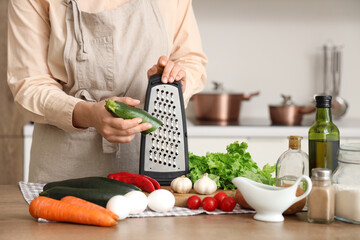Woman grating zucchini at table in kitchen, closeup