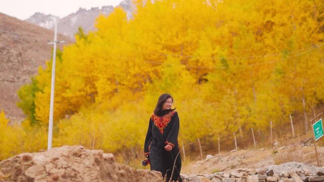 Indian woman holding camera in hand and walking in autumn trees background at Padum, Zanskar, India. Tourist wearing traditional dress Phiran. Girl in the mountains with fall background.