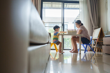 Asian mother is drawing with a child boy with colored pencil at a table. The child is sitting in a yellow chair