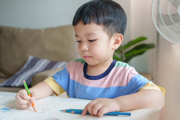Asian boy is sitting at a table and coloring with colored pencil on paper. Concept of creativity and playfulness