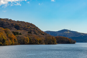 Autumn foliage paints the hillsides around the lake ; A serene lake surrounded by colorful autumn trees ; A breathtaking view of Lake Ashi in autumn