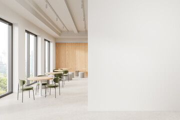 Modern cafe interior with wooden tables, green chairs, large windows, and a blank white wall for...