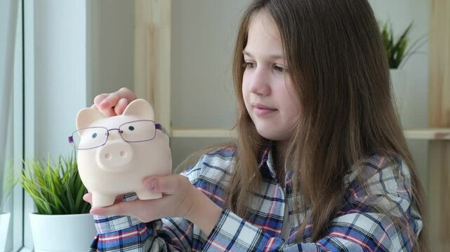 Cute smiling preteen girl holding pink piggy bank in glasses and putting coins in it. Child saving money, family savings concept