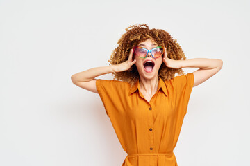 Happy woman with curly hair in sunglasses expressing joy, wearing an orange shirt against a plain white background, showcasing excitement and positivity in lifestyle imagery