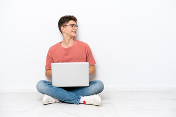 Teenager Russian man holding pc sitting on the floor isolated on white background laughing in lateral position