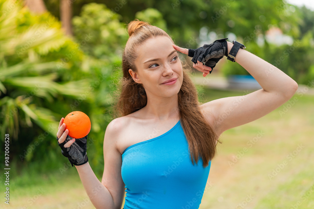 Poster Young pretty sport girl holding an orange at outdoors having doubts and with confuse face expression