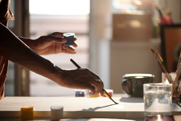 Hand grabbing paintbrush in bright studio with various art supplies scattered on desk. Sunlight...