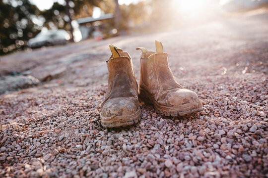 A pair of worn brown boots placed on a ground covered with small pebbles.