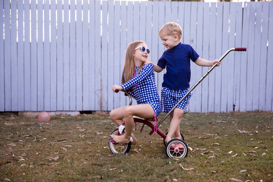 siblings smiling at each other while playing on a trike in the backyard