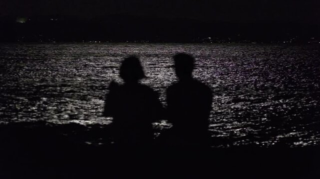 満月に光る海を眺めながら話す男と女 A man and a woman talking on the beach at night under the moonlight