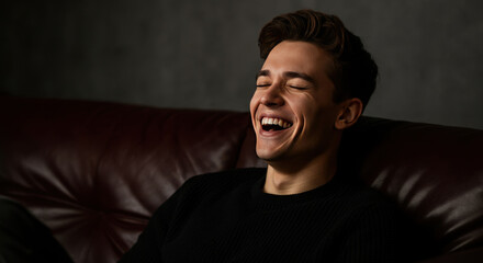 Fototapeta premium Smiling young man laughing on a leather couch. Indoor portrait with dark background