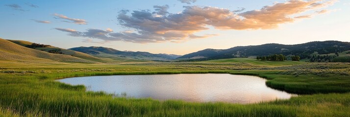 Fototapeta premium A tranquil pond in a field of green grass Mountains loom in the backdrop Clouds scatter the sky