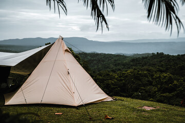 Obraz premium A triangle-shaped tent is pitched on a grassy hill, overlooking a lush green forest and distant mountains under a cloudy sky, creating a serene and scenic camping atmosphere.