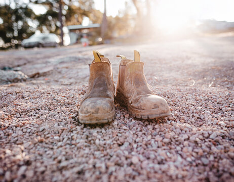A pair of worn brown boots placed on a ground covered with small pebbles.