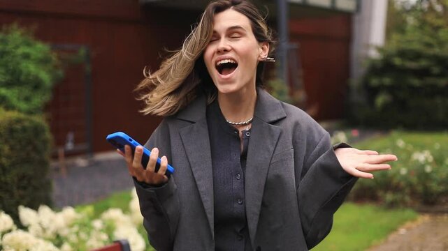 Attractive blonde woman look surprised wow hold using mobile cell phone. Girl look on smartphone and doing winner gesture. Girl just found out big win news, point on phone, wow, walking on the street.