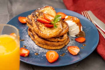 French toast garnished with strawberries and mint leaves with ice cream drizzled with caramel sauce and powdered sugar on a blue plate accompanied by a glass of orange juice on a dark background