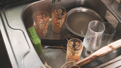 Untidy metal sink full of dirty dishes and tableware. Pile of dirty washing up utensils in the kitchen.