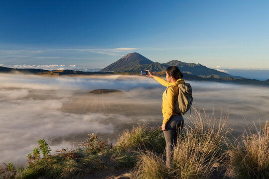 Asia, Indonesia, Java, Bromo Semeru national park, mature female tourist trekking at mount Bromo at sunrise, standing at viewpoint using mobile phone to take photographs

