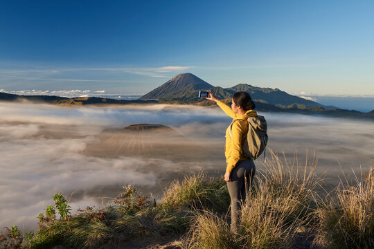 Asia, Indonesia, Java, Bromo Semeru national park, mature female tourist trekking at mount Bromo at sunrise, standing at viewpoint using mobile phone to take photographs

