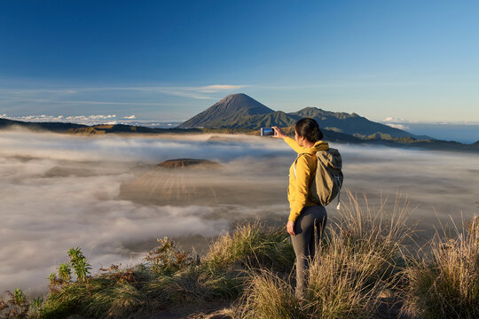 Asia, Indonesia, Java, Bromo Semeru national park, mature female tourist trekking at mount Bromo at sunrise, standing at viewpoint using mobile phone to take photographs

