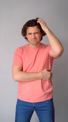 Confused man thinks scratching head by hand looking at camera on gray background. Curly middle aged guy feeling misunderstanding, trying to figure out situation. Pensive male wear jeans pink t-shirt.