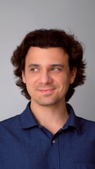 Portrait of man looking by side with sly tricky face on gray background. Curly middle aged guy having tricky plan in mind with smile. Wearing blue shirt. Showing emotions through facial expressions.