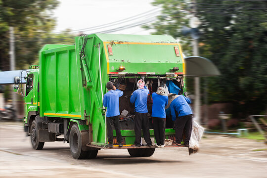 Garbage truck is running on the road to collect garbage with a garbage collector standing at the back of the truck.