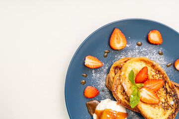 French toast garnished with strawberries and mint, with ice cream drizzled with caramel sauce, on a blue plate and orange juice on a white plate. Place for text.