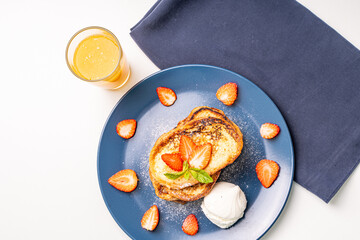 French toast decorated with strawberries and mint leaves with ice cream drizzled with caramel sauce and powdered sugar on a blue plate and a glass of orange juice on a white background, table