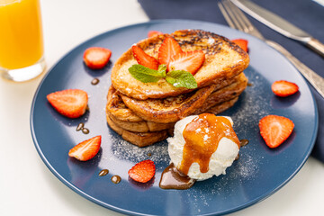French toast decorated with strawberries and mint leaves with ice cream drizzled with caramel sauce and powdered sugar on a blue plate and a glass of orange juice on a white background, table
