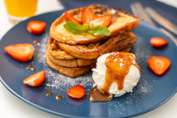 French toast decorated with strawberries and mint leaves with ice cream drizzled with caramel sauce and powdered sugar on a blue plate and a glass of orange juice on a white background, table