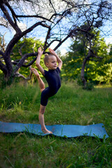 Fototapeta premium little 5-year-old girl in a black minimalist overall does gymnastics in the garden, outdoors