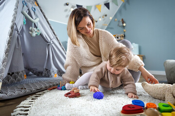Mom plays with her little child in a kid room with toys and stuffed animals, a childrens wigwam tent in the background © leszekglasner