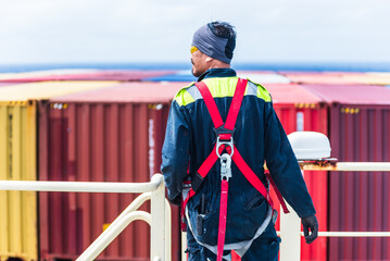 Standing on the deck of a massive container ship, a focused sailor secures his safety harness,...