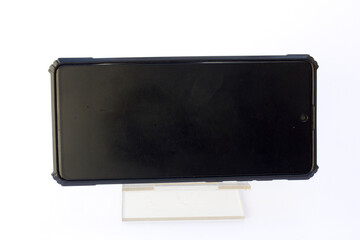 A modern smartphone with a black case mounted on a white stand against a plain, light background....