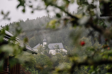 Bavaria, Germany - September 14, 2024 - A misty view of a Bavarian church with onion domes nestled in a forest, framed by foliage and rooflines on a rainy day..