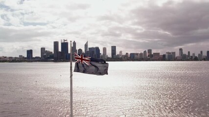 A flagpole with an Australian flag is waving in the wind. Behind the flagpole, a river flows to the skyline of Perth, Australia - Powered by Adobe