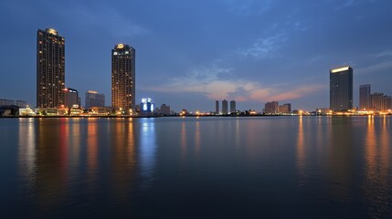 Naklejka premium City skyline at dusk with high-rise buildings illuminated, reflecting on the calm water, under a deep blue and cloudy sky.