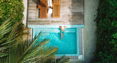 A young woman in a hat and swimsuit is relaxing in the clear water of a private pool. Top view of...