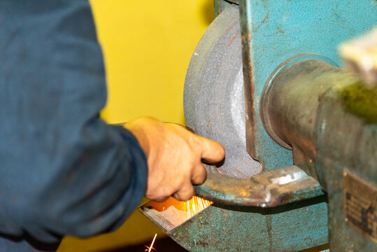 Close-up of a crew member operating a stationary grinder aboard a merchant vessel, focusing on the worker&rsquo;s hands, the workpiece, and the spinning grinding wheel in action.